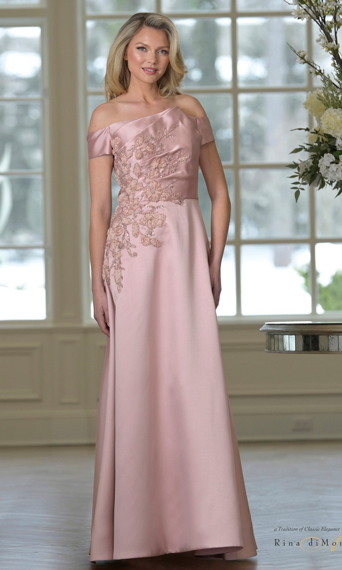 A woman stands indoors before large windows, wearing the Rina Di Montella RD3501 long formal dress in deep rose, featuring an off-shoulder floral embroidered bodice and an elegant a-line skirt.